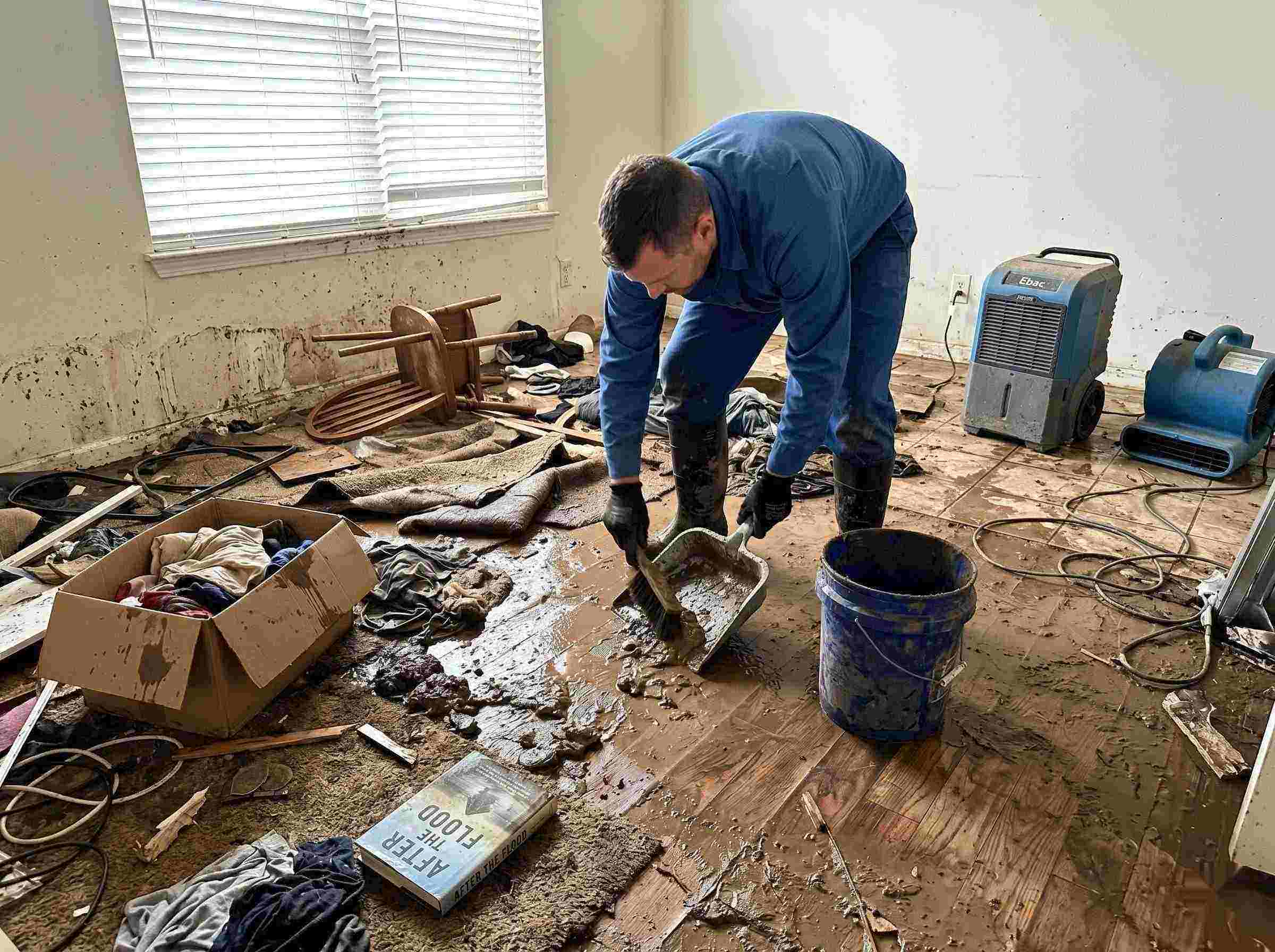 Realistic photo of a technician performing mud and debris removal after a home flood in Rochester, showing soaked furniture, wet flooring, and scattered debris during post-flood cleanup.